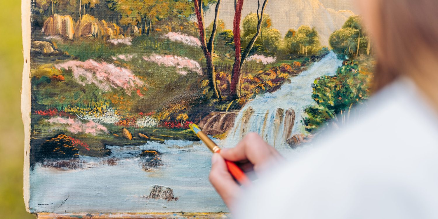 Selective focus on a hand painting with a brush on a canvas with a painting of a forest in a park